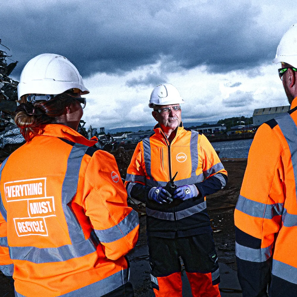 Three people in high visibility jackets and hard hats standing near a body of water.