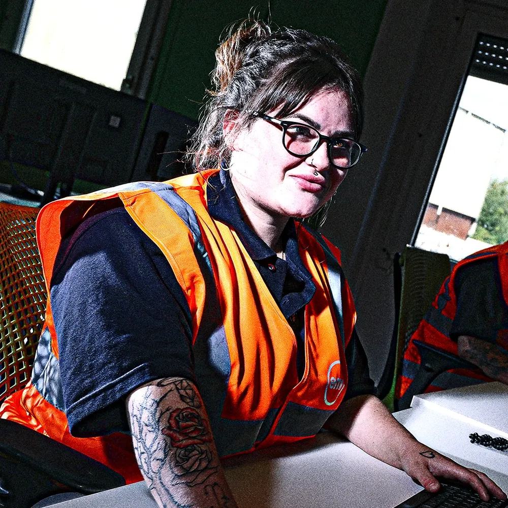 A person wearing an orange safety vest and glasses sitting at a desk and using a laptop.